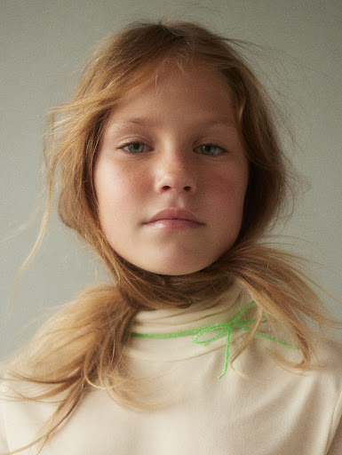 Close-up portrait of a fair-skinned child with long blonde hair, wearing a cream-colored turtleneck sweater with neon green embroidery.