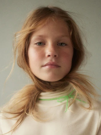 Close-up portrait of a fair-skinned child with long blonde hair, wearing a cream-colored turtleneck sweater with neon green embroidery.