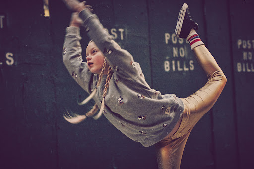 A young gymnast in gray sweatshirt and gold leggings performs a backbend with one leg extended up against a green background indoors.