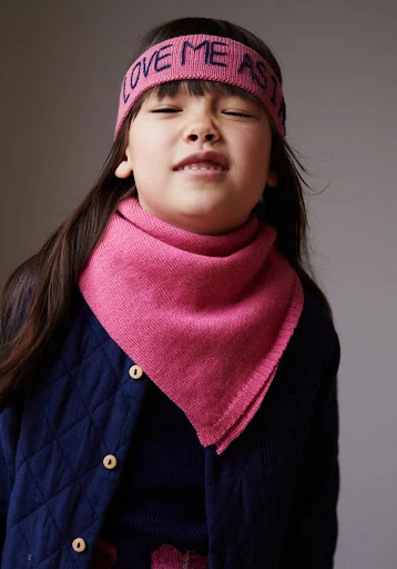 A young child wearing a pink headband and scarf, closes their eyes and makes a facial expression in a studio setting.