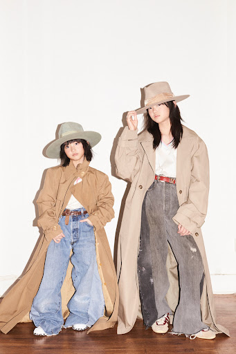 Two stylish young women in wide-brimmed hats and trench coats pose against a white wall, showcasing a vintage-inspired fashion look.