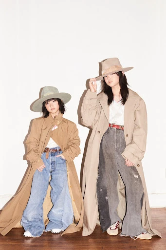 Two stylish young women in wide-brimmed hats and trench coats pose against a white wall, showcasing a vintage-inspired fashion look.