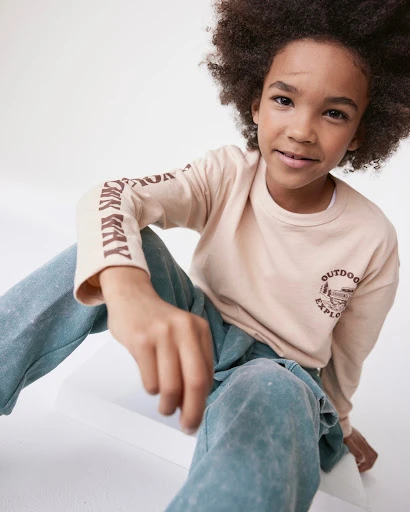 Portrait of a young kid with an afro hairstyle, wearing a beige long-sleeved shirt and blue pants while sitting indoors.