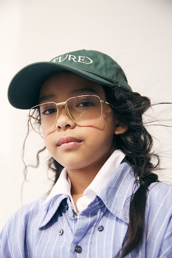 Young girl with long dark hair, wearing a green baseball cap and large gold-rimmed glasses, against a white background. Fashion portrait.