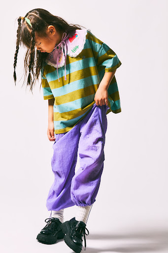 Full-body shot of a young girl in a striped shirt and purple pants, posing on a white background while looking down and to the left.