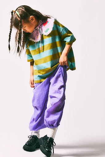 Full-body shot of a young girl in a striped shirt and purple pants, posing on a white background while looking down and to the left.