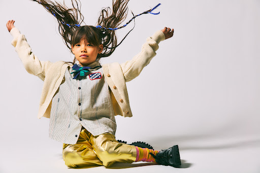 Young child with long hair flies upward in a photo studio. The child wears gold pants, a striped shirt, and a cream-colored cardigan.