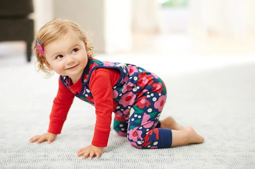 Toddler in a floral print outfit crawls on a rug in a brightly lit room, looking up and smiling with a pink butterfly clip in her hair.