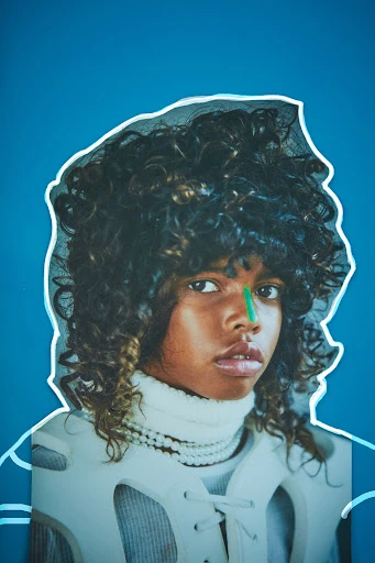 Striking portrait of a young person with curly hair and a green stripe on their nose, wearing a white top, against a blue background.