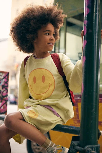 Child with afro sits outdoors in a yellow smiley-face sweatshirt and shorts set with a red backpack, smiling and looking to the side.