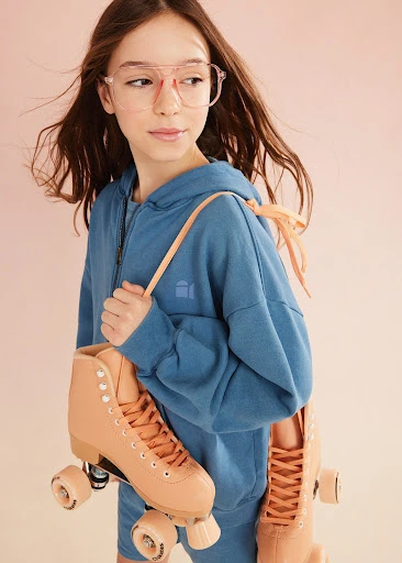 Thora, a blonde-haired model, poses in a photo studio against a blue background. She is wearing a light blue knit top.