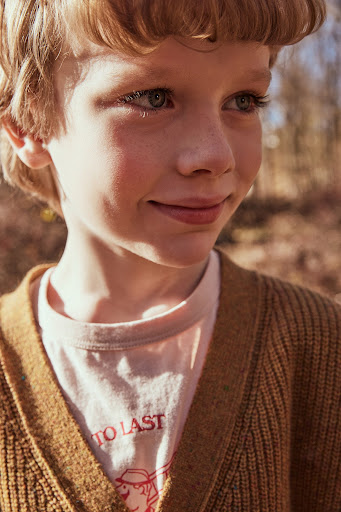 Close-up portrait of a smiling young boy with blond hair and blue eyes, wearing a tan cardigan in a natural, outdoor setting.