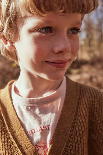 Close-up portrait of a smiling young boy with blond hair and blue eyes, wearing a tan cardigan in a natural, outdoor setting.