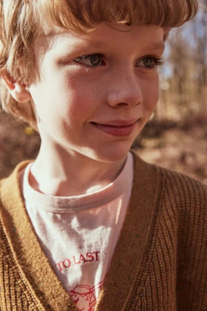 Close-up portrait of a smiling young boy with blond hair and blue eyes, wearing a tan cardigan in a natural, outdoor setting.