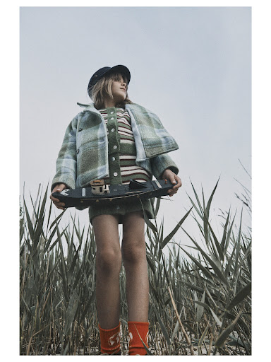Young girl in a plaid coat and skirt stands in a grassy field, holding a model sailboat, wearing a hat and red socks, shot from low angle.