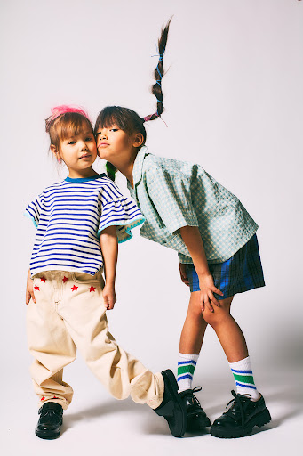 Two young girls in casual outfits pose in a studio setting, one with a ponytail and the other with pink hair. Smiling, they are captured in a playful moment.