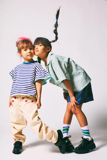 Two young girls in casual outfits pose in a studio setting, one with a ponytail and the other with pink hair. Smiling, they are captured in a playful moment.