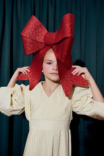 Girl poses indoors against green curtain wearing cream-colored dress and oversized red glittery bow headpiece, medium shot.