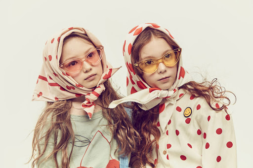 Two young girls wearing polka dot headscarves and colorful sunglasses pose in a studio shot on a white background.