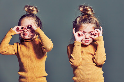 Two cute little girls with hair in a bun, smiling and posing in a photo studio wearing mustard yellow turtlenecks on grey background.