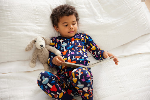 Young child lying on a white bed reading a book next to a stuffed dog toy, wearing mushroom-patterned pajamas in a cozy indoor setting.