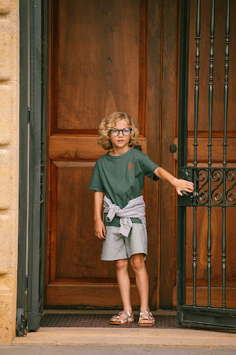 Young blonde child stands by a dark wood door with a metal gate, wearing an olive t-shirt, grey shorts, and sandals in an outdoor setting.