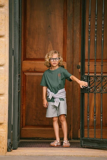 Young blonde child stands by a dark wood door with a metal gate, wearing an olive t-shirt, grey shorts, and sandals in an outdoor setting.