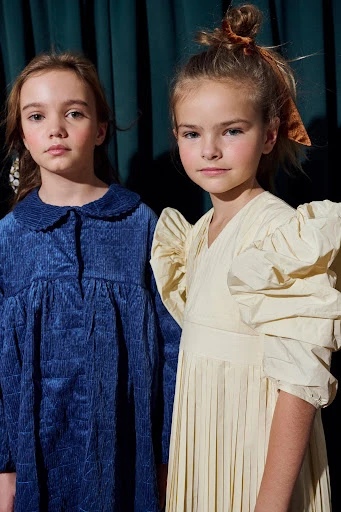 Two young girls pose indoors against a dark teal curtain backdrop, wearing textured dresses in blue and off-white, looking at the camera.
