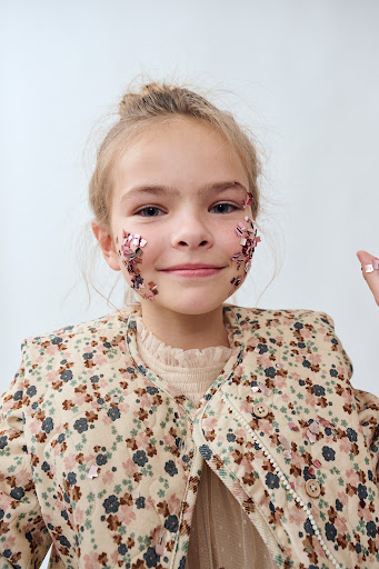 Young girl with hair in a bun smiles with pink glitter on her face, wearing a floral print jacket in a studio shot.