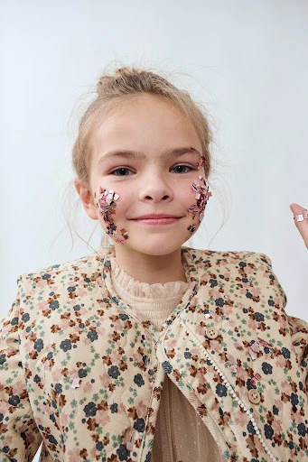 Young girl with hair in a bun smiles with pink glitter on her face, wearing a floral print jacket in a studio shot.