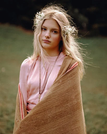 Blonde woman with a flower crown wrapped in a brown blanket stands outdoors against a soft green background in a portrait style.