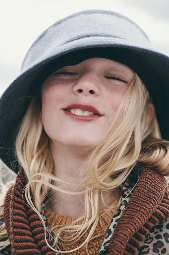 Portrait of smiling young girl with eyes closed wearing a gray hat and brown patterned sweater with a scarf, outdoors on an overcast day.