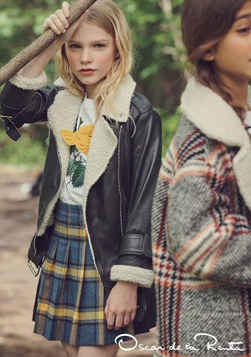 Two young girls wearing outerwear stand outdoors; one in a plaid jacket, the other in a shearling-lined jacket and plaid skirt.