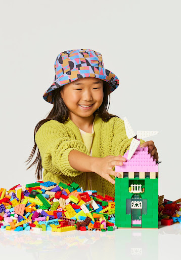Smiling Asian child in patterned hat builds a castle from colorful plastic toy blocks in a light studio setting.