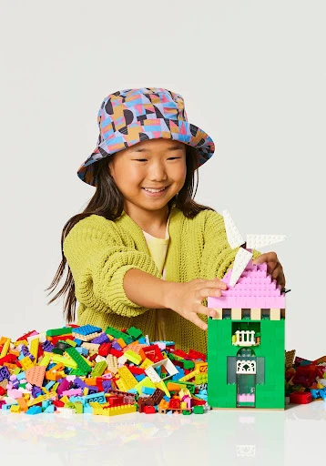 Smiling Asian child in patterned hat builds a castle from colorful plastic toy blocks in a light studio setting.