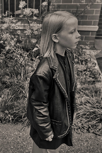 Black-and-white portrait of a child in a studded black jacket, viewed in profile, standing outdoors amid greenery and a brick wall.