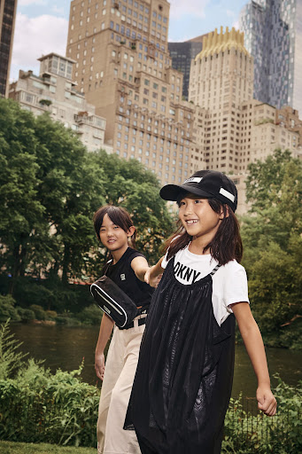 Two smiling young girls in stylish black and white outfits walk in an urban park setting with tall buildings in the background.