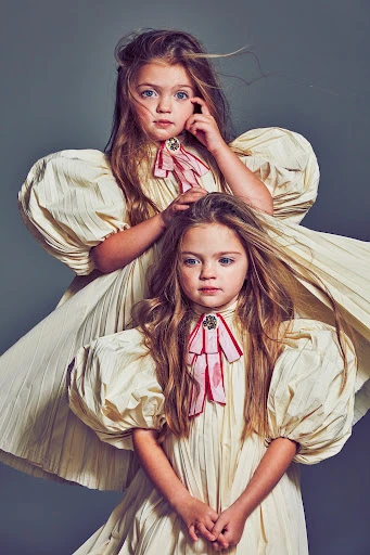 Two young girls in matching cream-colored dresses with puffy sleeves pose indoors against a gray backdrop, looking directly at the camera.