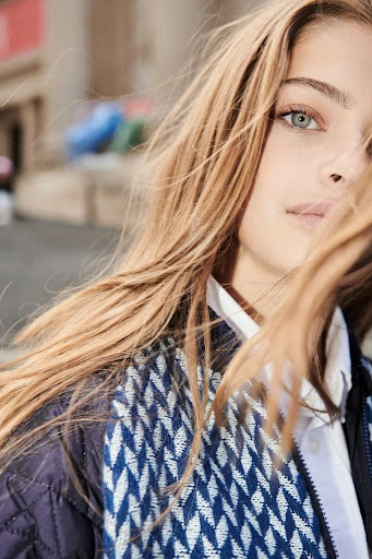 Close-up portrait of a young adult female model with blonde hair in an outdoor setting, wearing a patterned jacket and collared shirt.