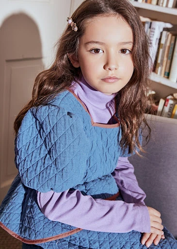 Young girl with long brown hair sits indoors wearing a blue quilted dress over a light purple turtleneck, styled with a hair clip.