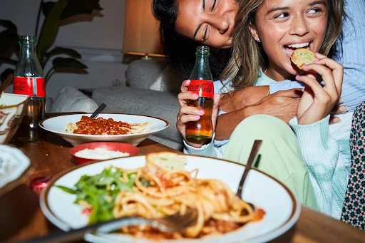 Two young women share a meal of spaghetti and garlic bread, enjoying Coca-Cola in a cozy indoor setting with warm lighting.