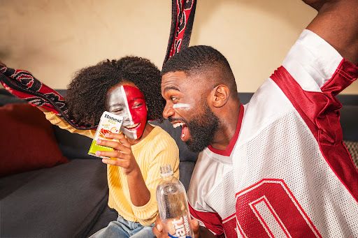 Enthusiastic father and daughter cheer while watching a game, faces painted, with sports drinks and a football jersey, indoor setting.