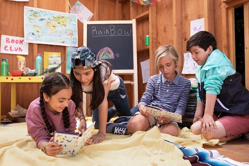 Four children are inside a wooden structure, reading books and papers on a yellow blanket, with artwork and school supplies nearby.
