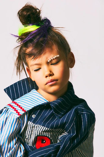 Portrait of a child model in a studio wearing a colorful oversized shirt and decorative pearls, with a neutral expression against white.