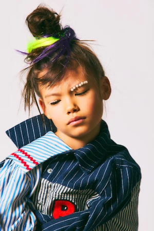 Portrait of a child model in a studio wearing a colorful oversized shirt and decorative pearls, with a neutral expression against white.