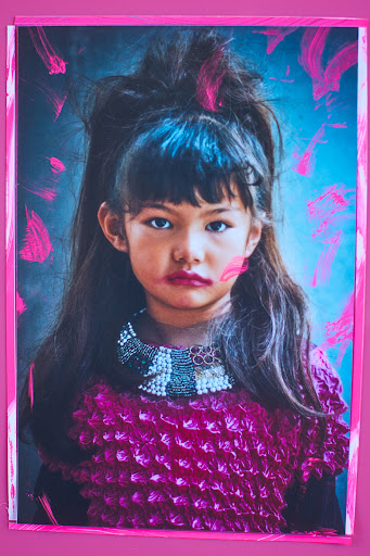 Portrait of a young girl with makeup and a magenta outfit against a pink frame and a soft blue backdrop with magenta paint strokes.