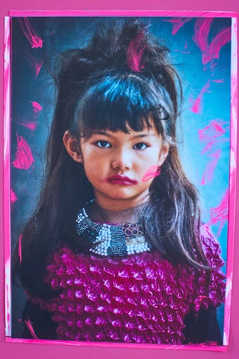 Portrait of a young girl with makeup and a magenta outfit against a pink frame and a soft blue backdrop with magenta paint strokes.