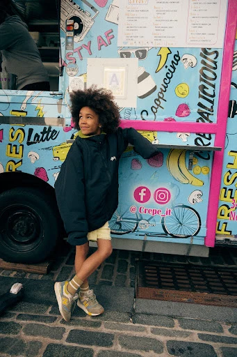 Young person leaning against a colorful food truck with a menu, wearing a dark jacket and shorts, standing on a brick surface in daylight.