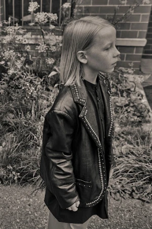 Monochrome portrait of a child wearing a studded black leather jacket outdoors near foliage.