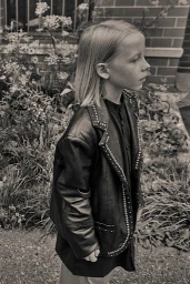 Monochrome portrait of a child wearing a studded black leather jacket outdoors near foliage.